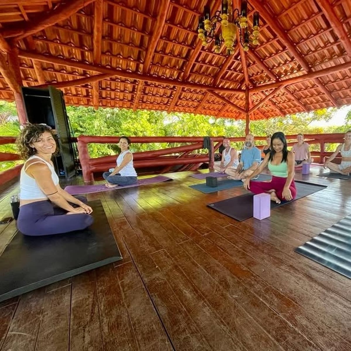 Small group seated in the open-air Treehouse yoga pavilion under a tiled roof, jungle just beyond the railings